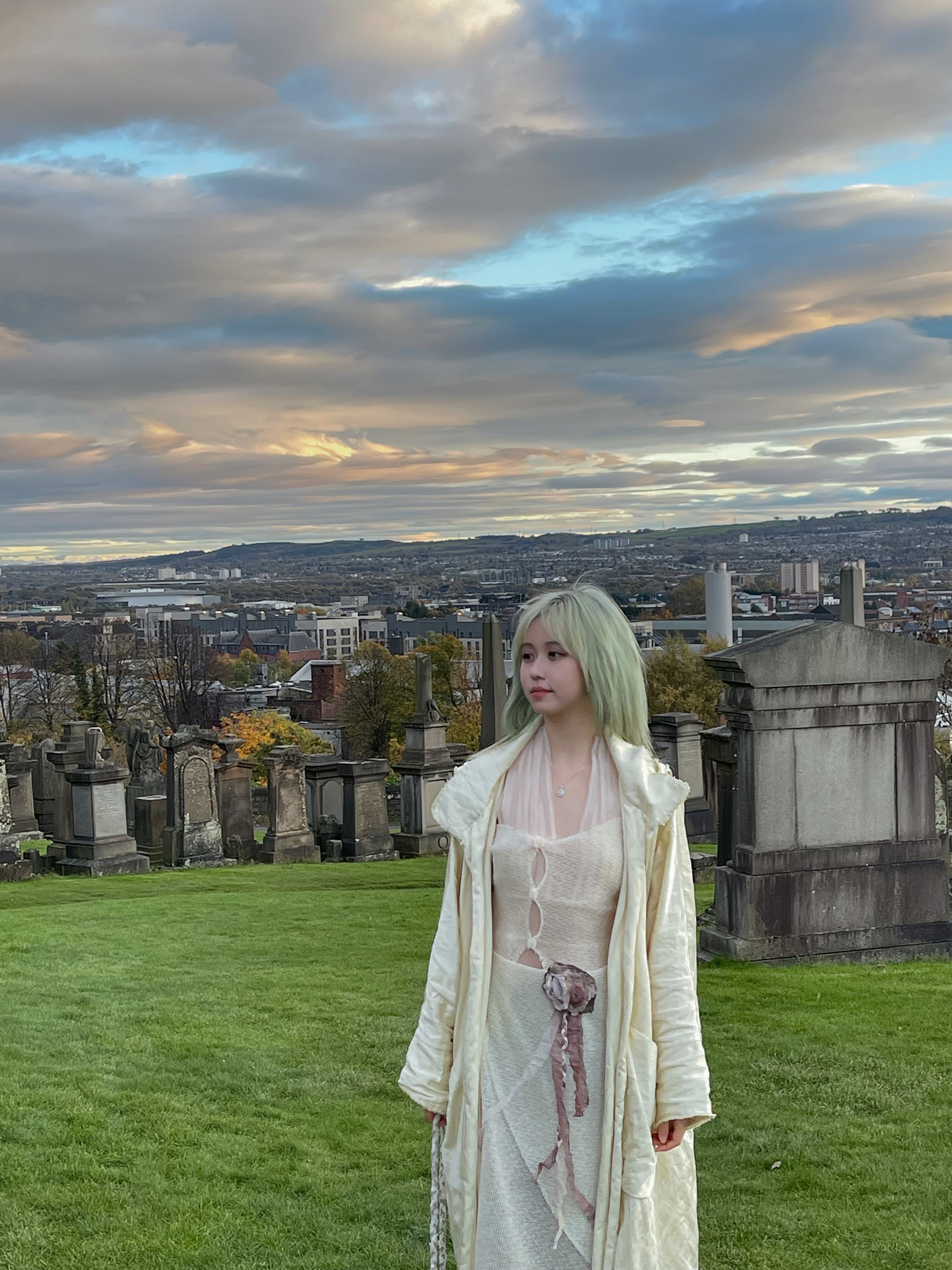 Violet standing among the monuments of the Glasgow Necropolis, with the city spread out behind under evening clouds