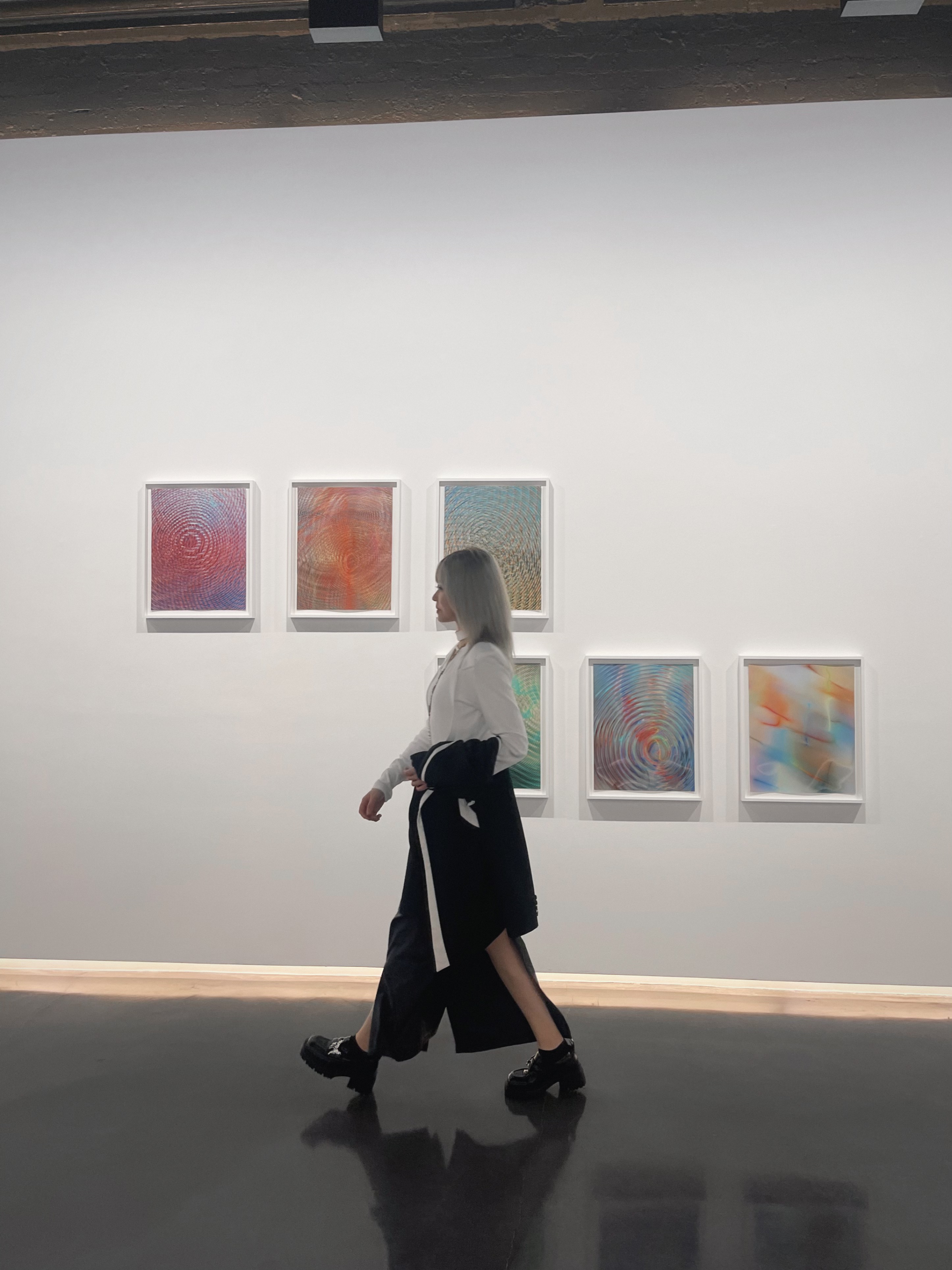 Violet walking past rows of small glowing colour prints on a white gallery wall in Mayfair