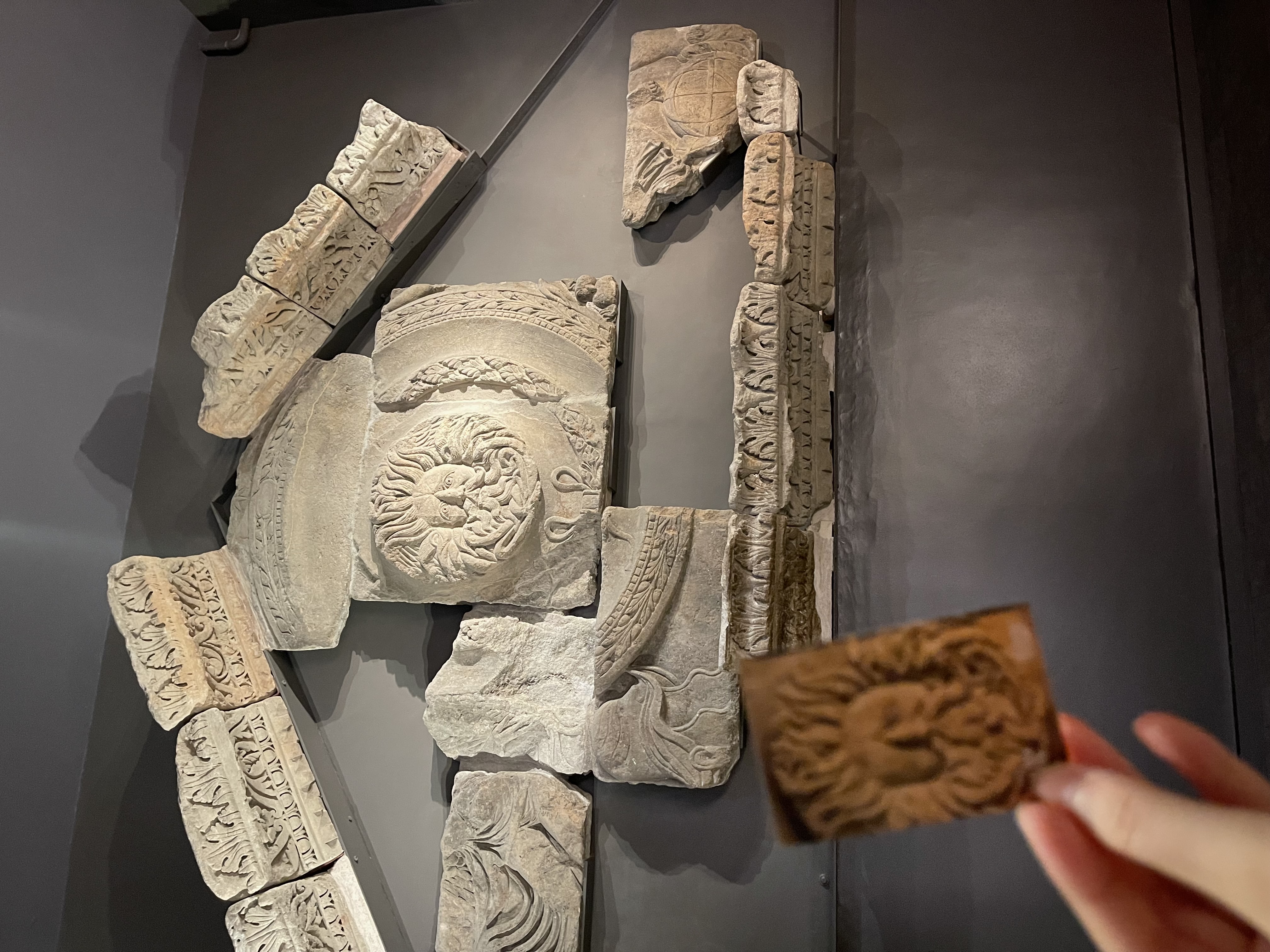 Carved Roman stone fragments mounted on a dark wall at the Roman Baths, with a hand holding up a small brown print of a carved face