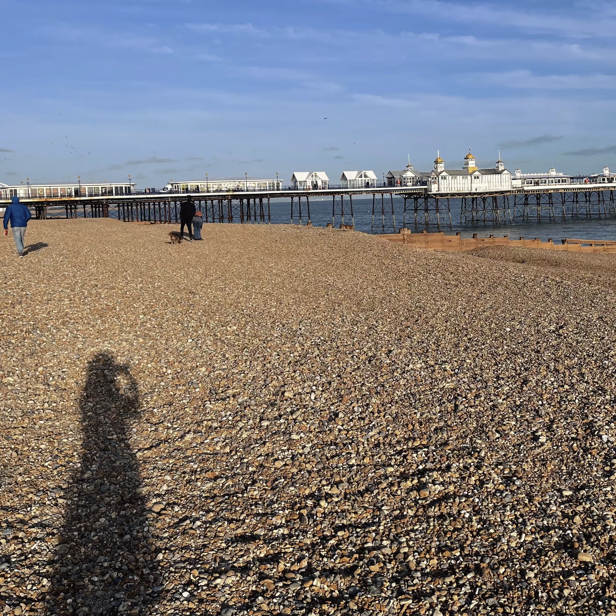 A long shadow of a person on the shingle beach at Eastbourne, with the pier in the distance