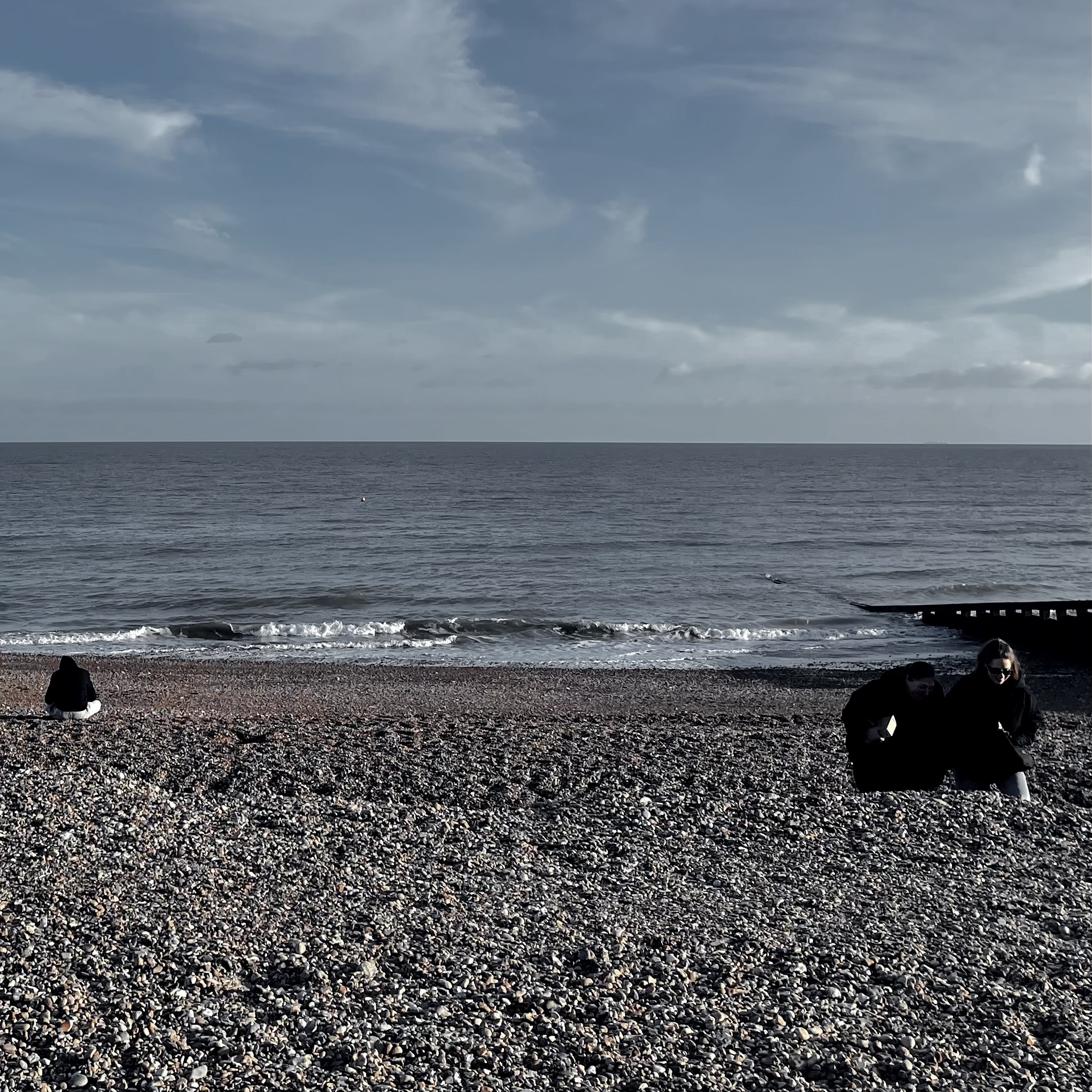 Waves on the shingle at Eastbourne under a pale sky, with two people sitting on the stones