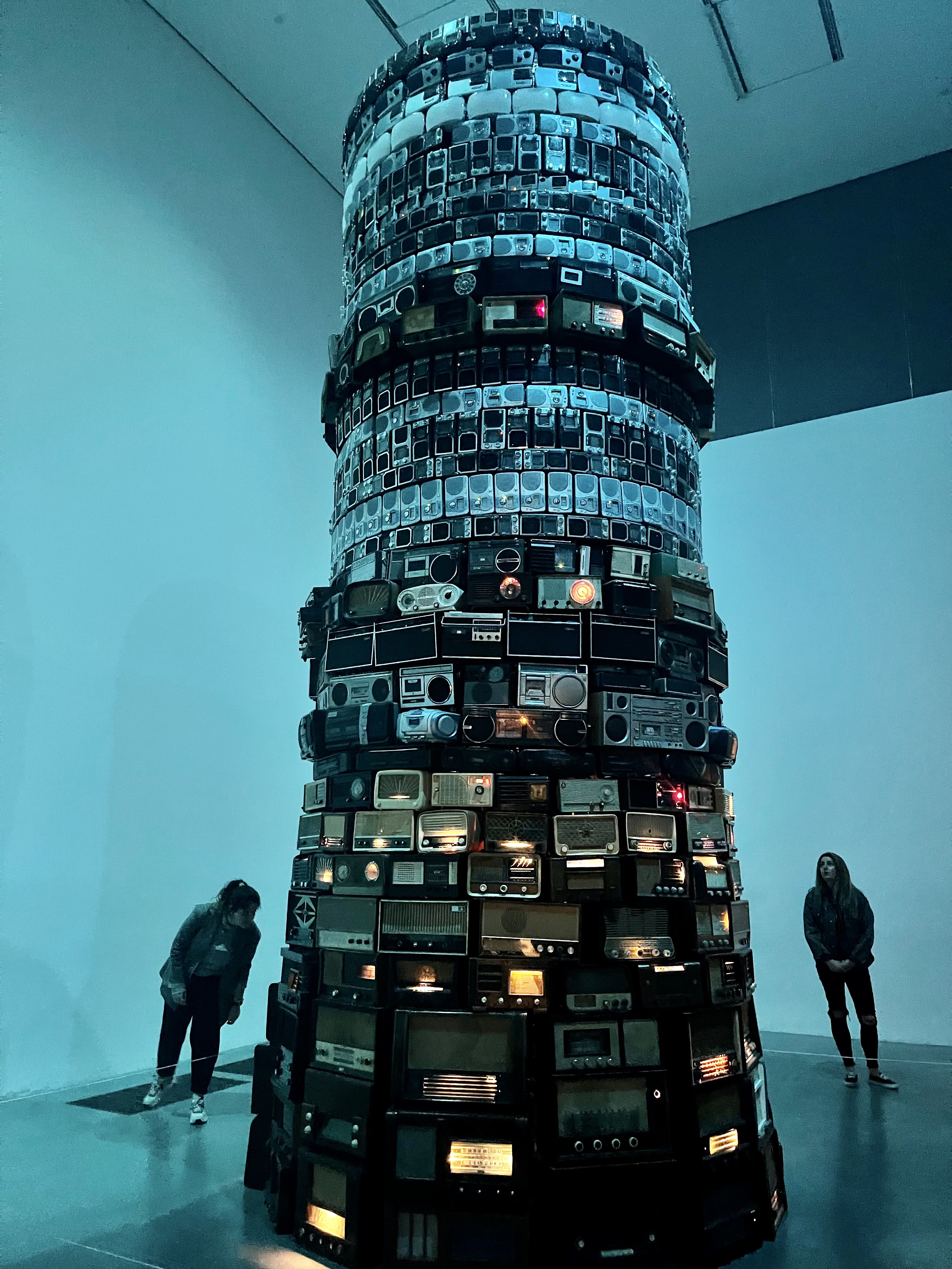 A tall tower of stacked old radios lit in blue at Tate Modern, with two visitors at its base