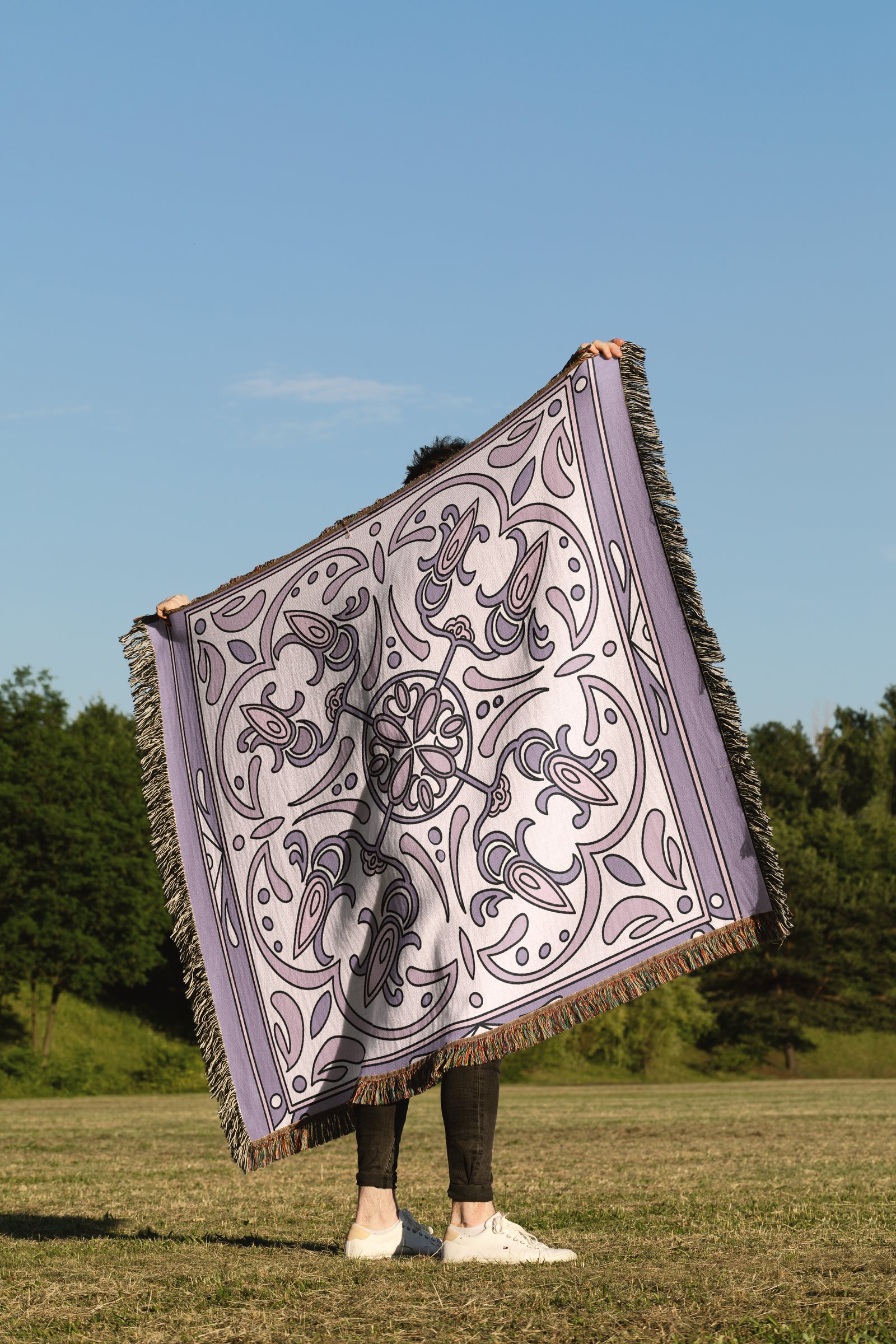 A person in a park holding up a large fringed woven throw printed with a lilac Ornata pattern